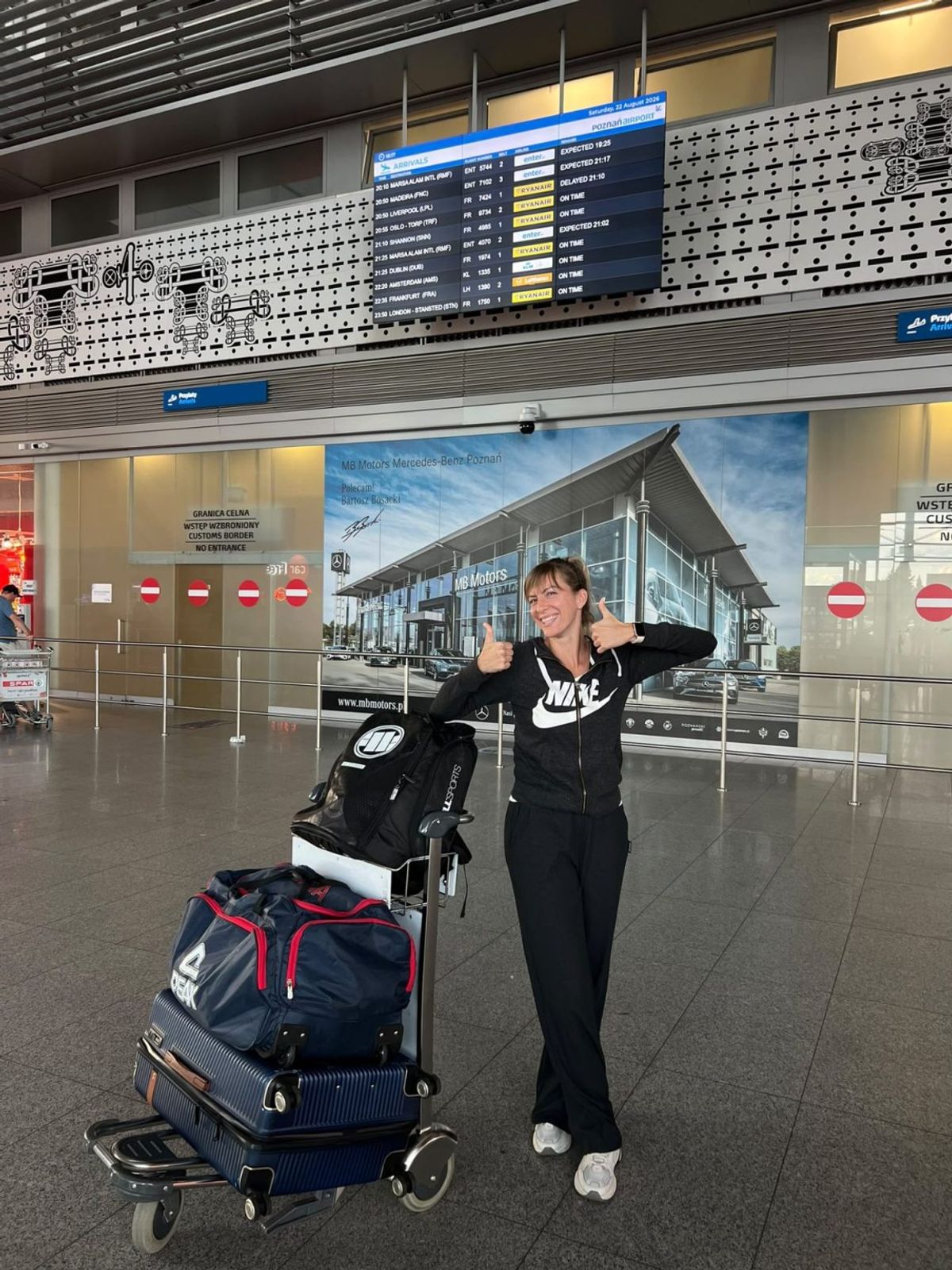 Jovana Popović w czarnym dresie z wózkiem bagażowym w hali przylotów lotniska, pokazuje kciuki w górę.