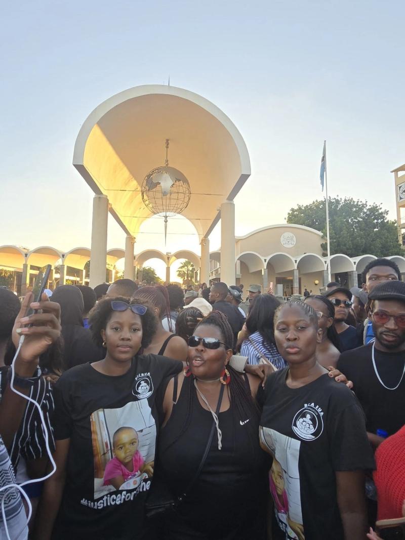 Re A Bua supporters in branded t-shirts gather outside Parliament at dusk.