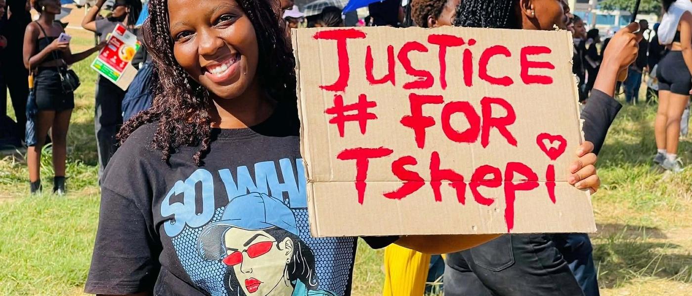 A young woman at the march holds a hand-painted 'Justice # for Tshepi' sign.