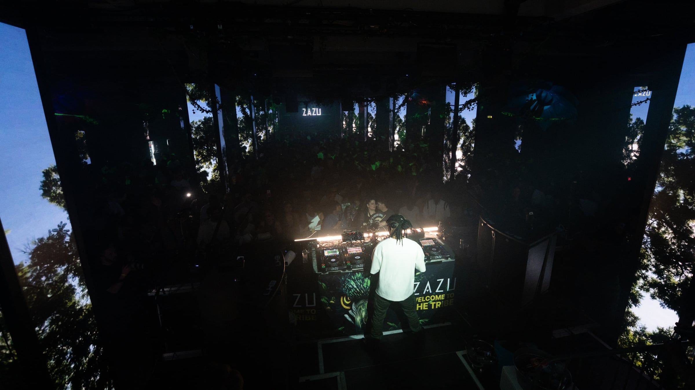 DJ in de booth van Zazu, met publiek en projecties van bomen