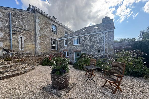 Gravel courtyard with wooden chairs, a small table and a stone planter beside stone buildings, with a dog on the steps