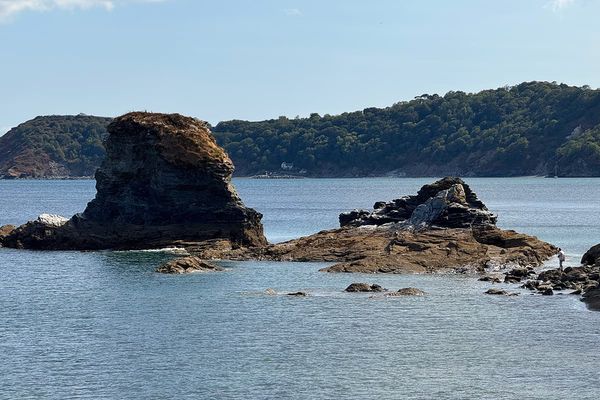 Rock stacks rising from calm blue sea, with a wooded headland and a small white house on the far shore
