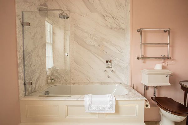 Deluxe bedroom bathroom with a marble-panelled bath and overhead shower, heated towel rail, pink walls and wooden toilet seat