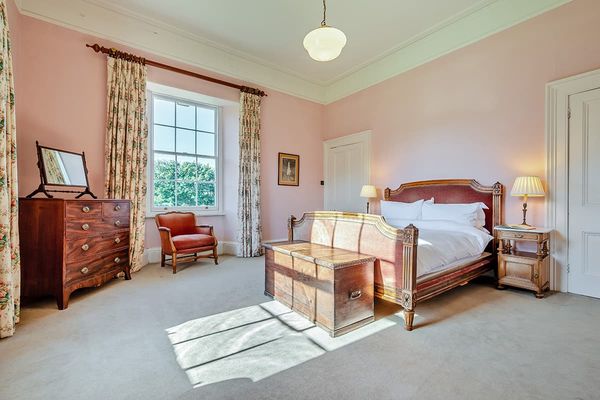 Standard bedroom with pink walls, a carved wooden bed, a chest at its foot and a red armchair by a sash window