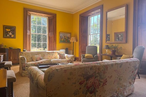 Sunlit living room with yellow walls, floral sofas, grey armchairs, large sash windows with wooden shutters and a tall mirror