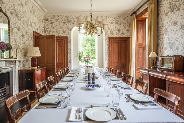 Formal dining room with a long table set with white linen, brass chandelier, floral wallpaper and a tall arched window