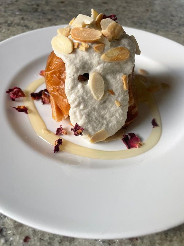 Baked apple topped with a thick cream, toasted flaked almonds and dried rose petals, with a swirl of sauce on a white plate