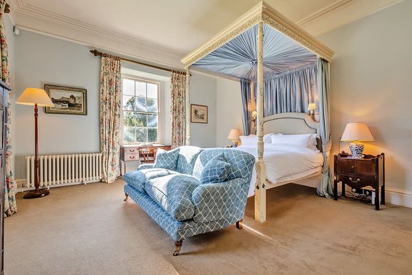 Deluxe bedroom with a painted four-poster bed and blue canopy, a blue patterned sofa, writing desk and sunlit sash window