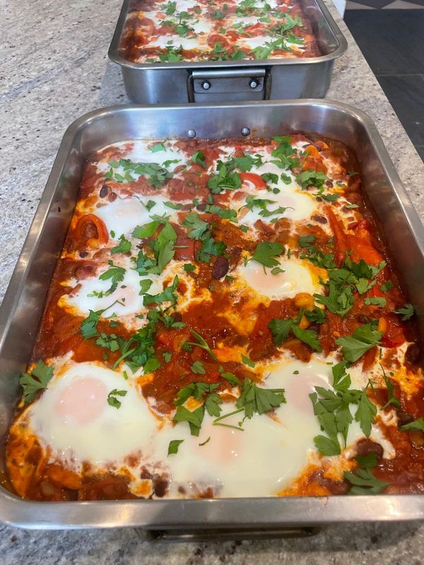 Two trays of eggs baked in a tomato, red pepper and bean sauce, scattered with fresh flat-leaf parsley