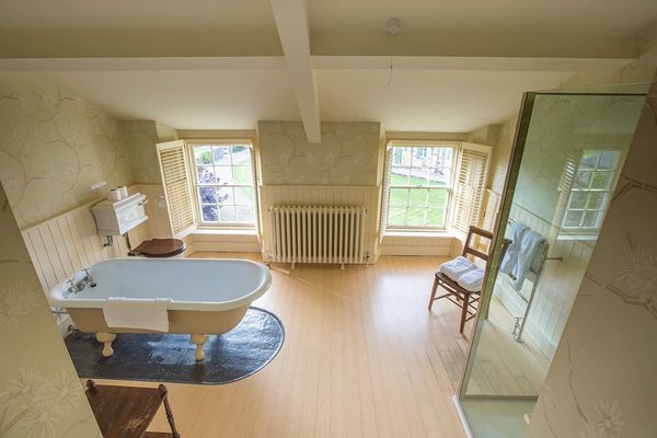 Spacious deluxe bedroom bathroom with a roll-top bath, glass shower, wooden floor and two sash windows overlooking a lawn