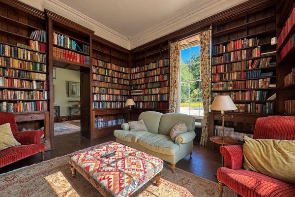 Library lined with wooden bookshelves, with a pale green sofa, red armchairs, a patterned footstool and a tall sash window
