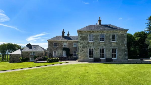 Tresillian House, a Georgian stone house with tall sash windows and slate roofs, behind a wide lawn under a clear blue sky
