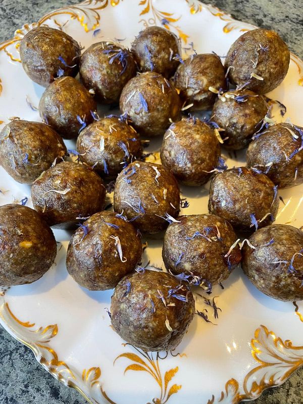 Round sweet energy balls scattered with dried blue flower petals on a white plate with gold scroll edging