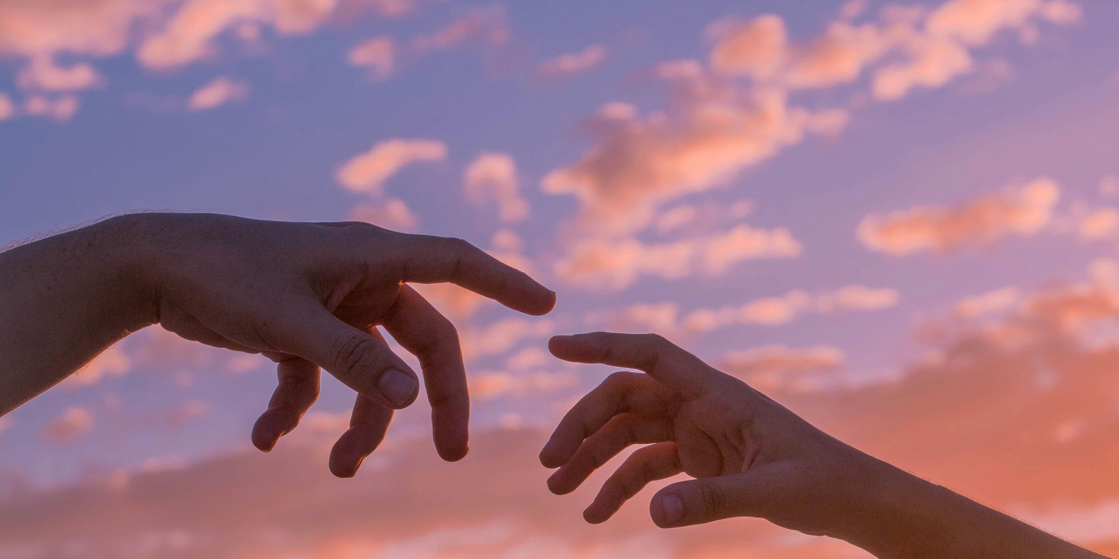 Reached out arms almost touching pointer fingers in front of a colorful clouded sky