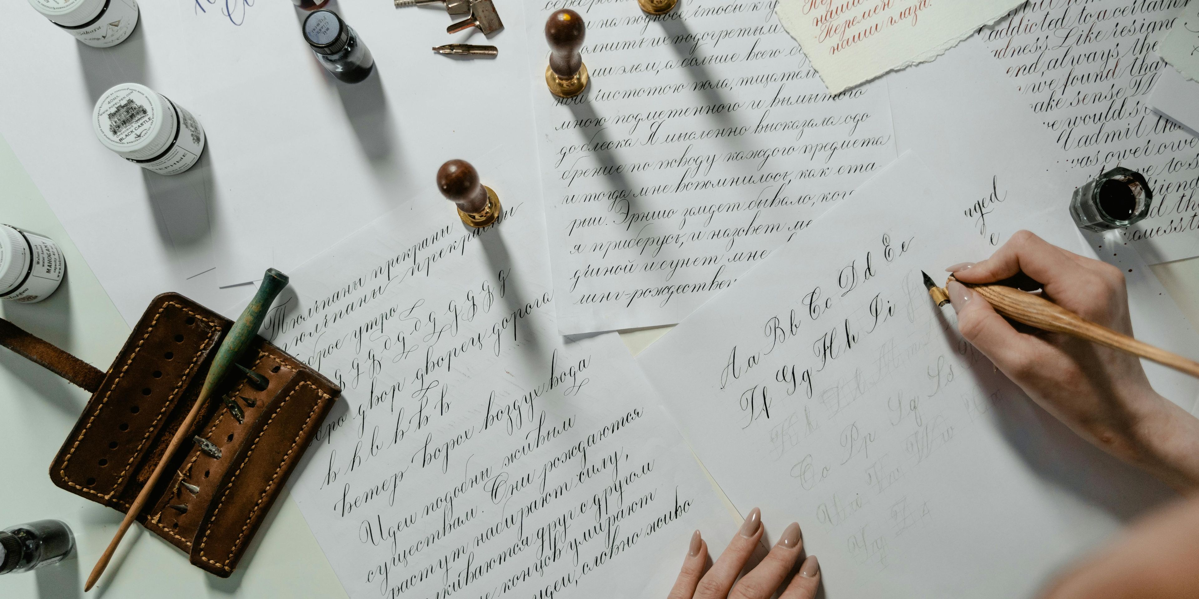 A ladies hands holding a quill, writing on a page, many previously written pages in the background.
