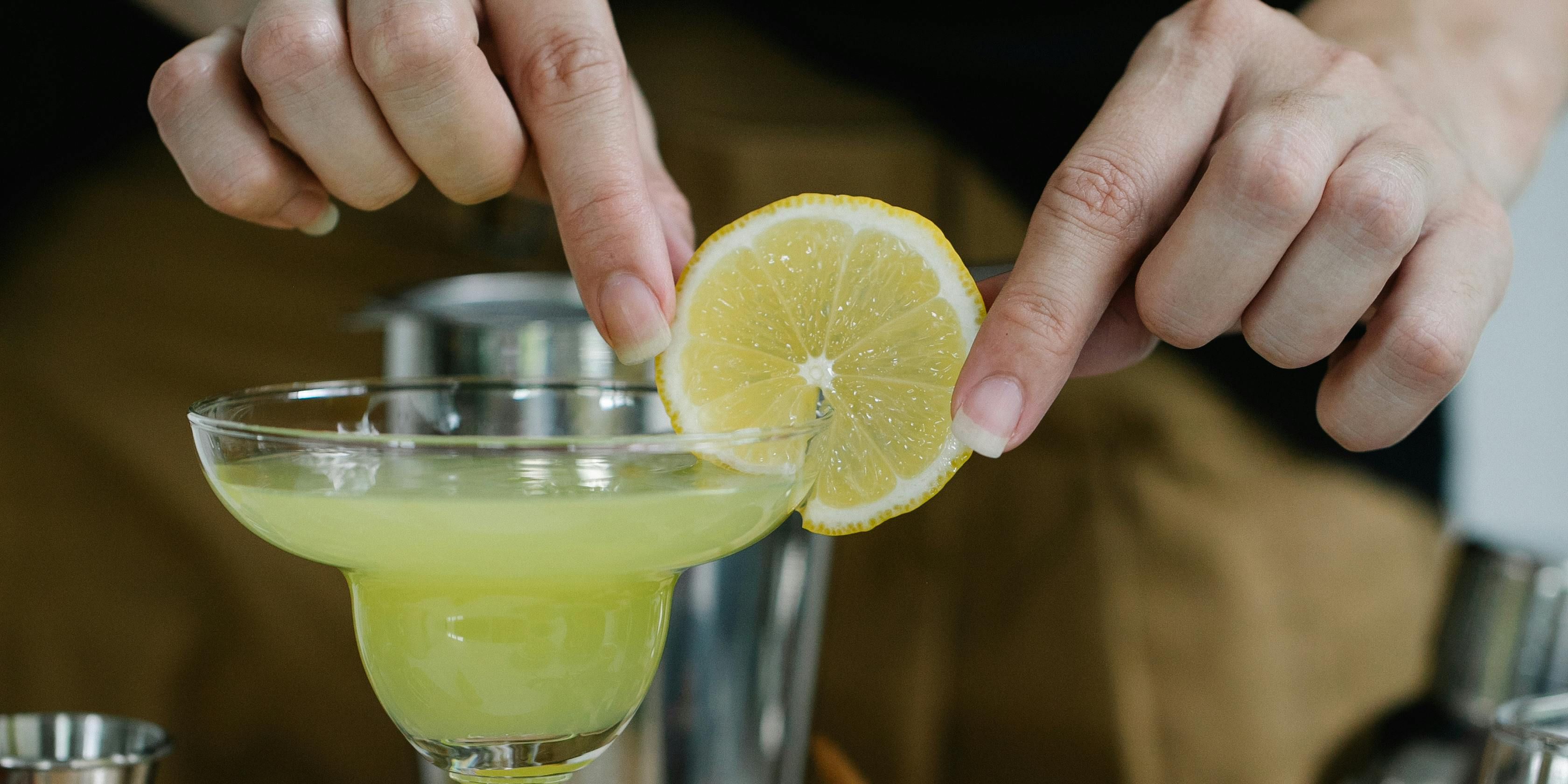 Person making a margarita finishing it off with a slice of lemon on the side of the glass