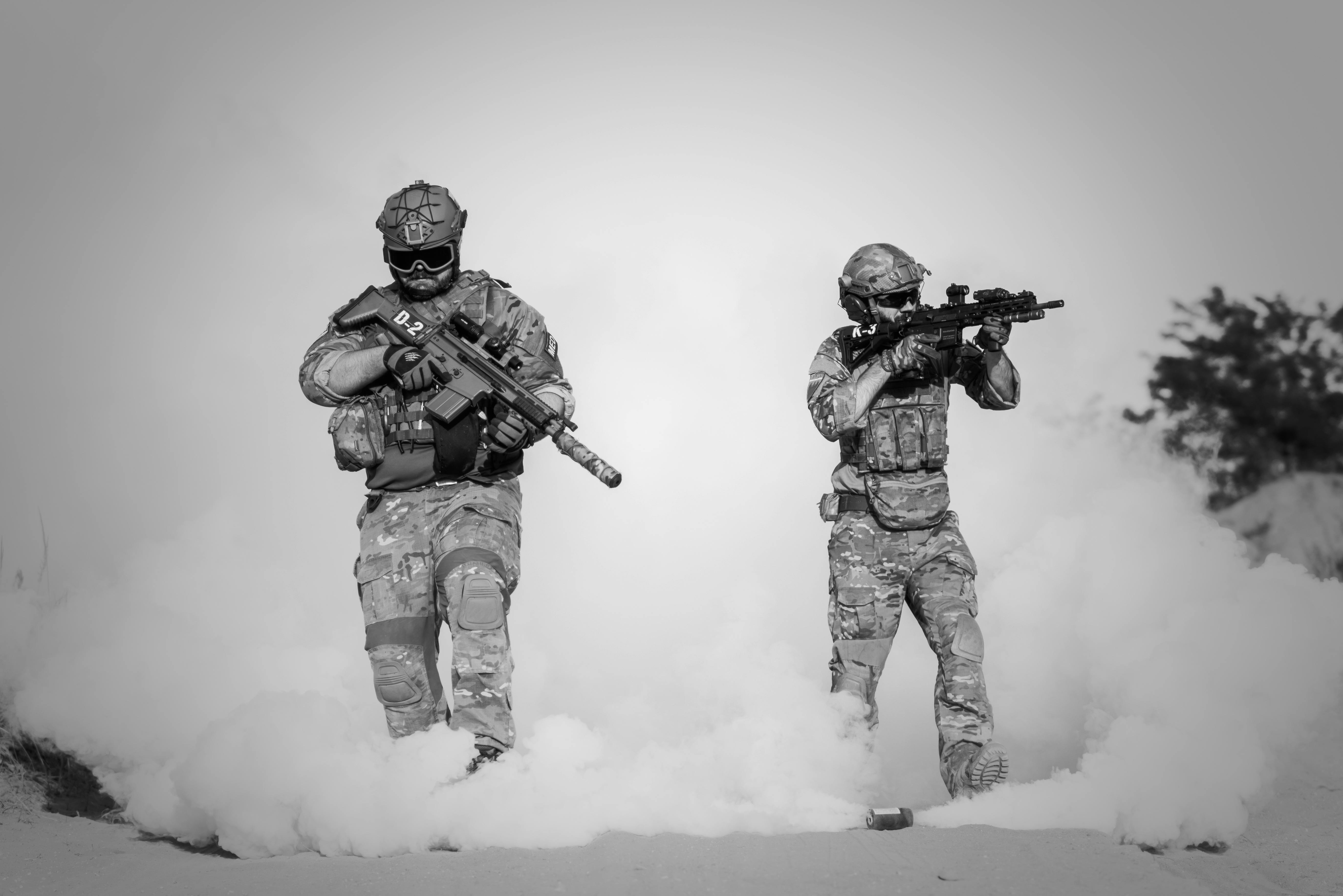 Two soldiers in camouflage tactical gear holding guns and walking through smoke grenades