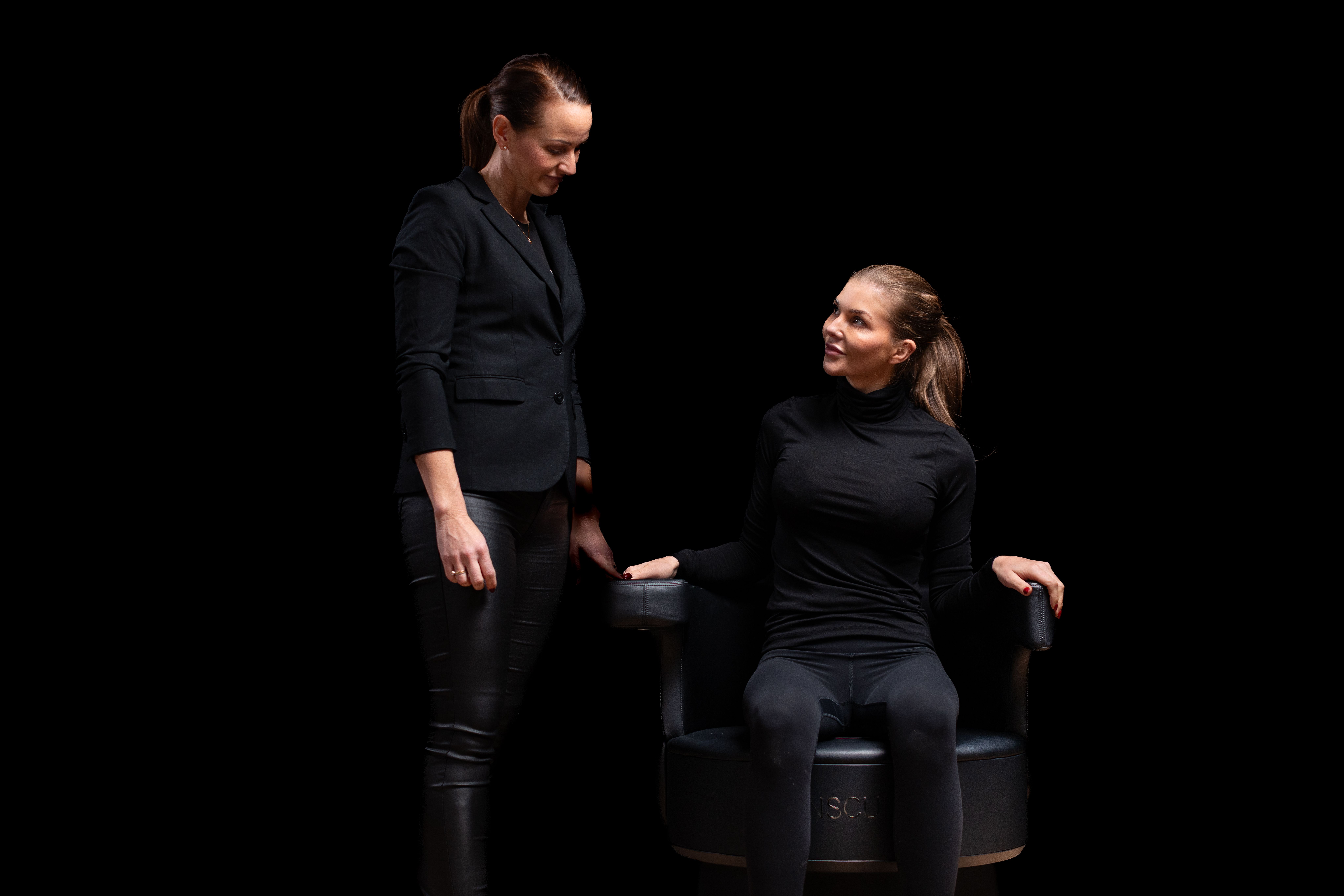 En person sitter på insculpt chair, en annen står på siden og passer på