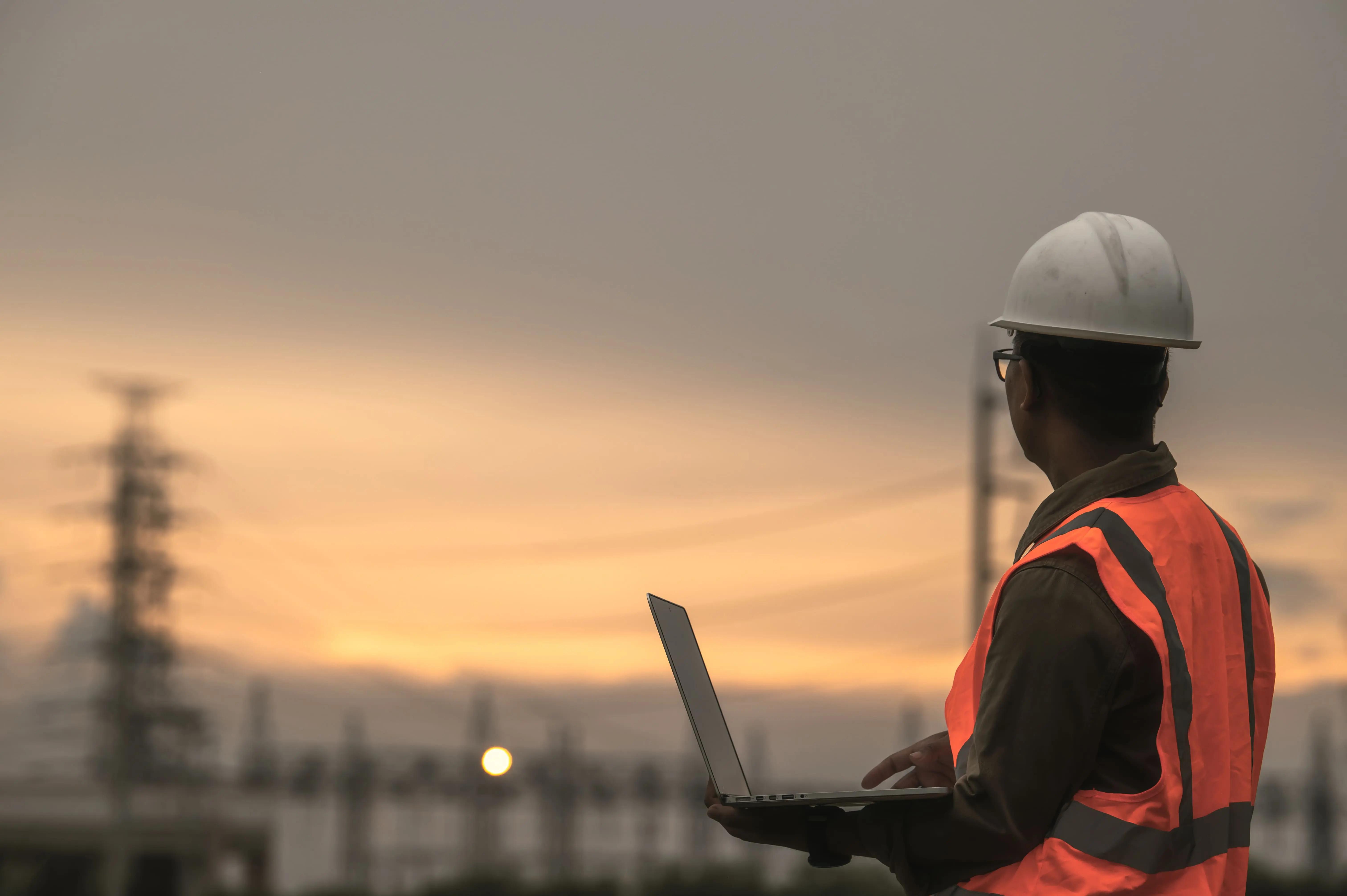 A man in a hard hat and orange vest is using a laptop computer