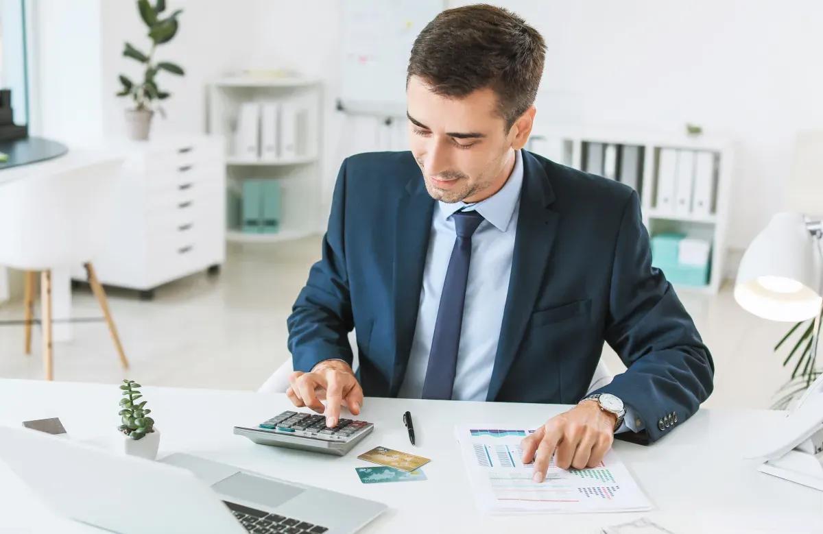 A man in a suit and tie is sitting at a desk using a calculator