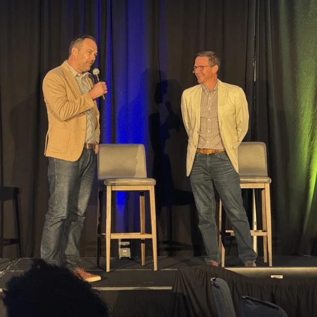 Two men are standing on a stage talking into microphones