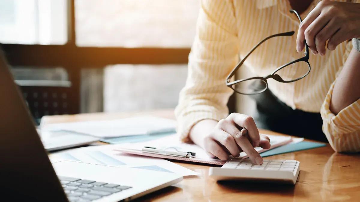 A woman is sitting at a desk using a calculator and a laptop