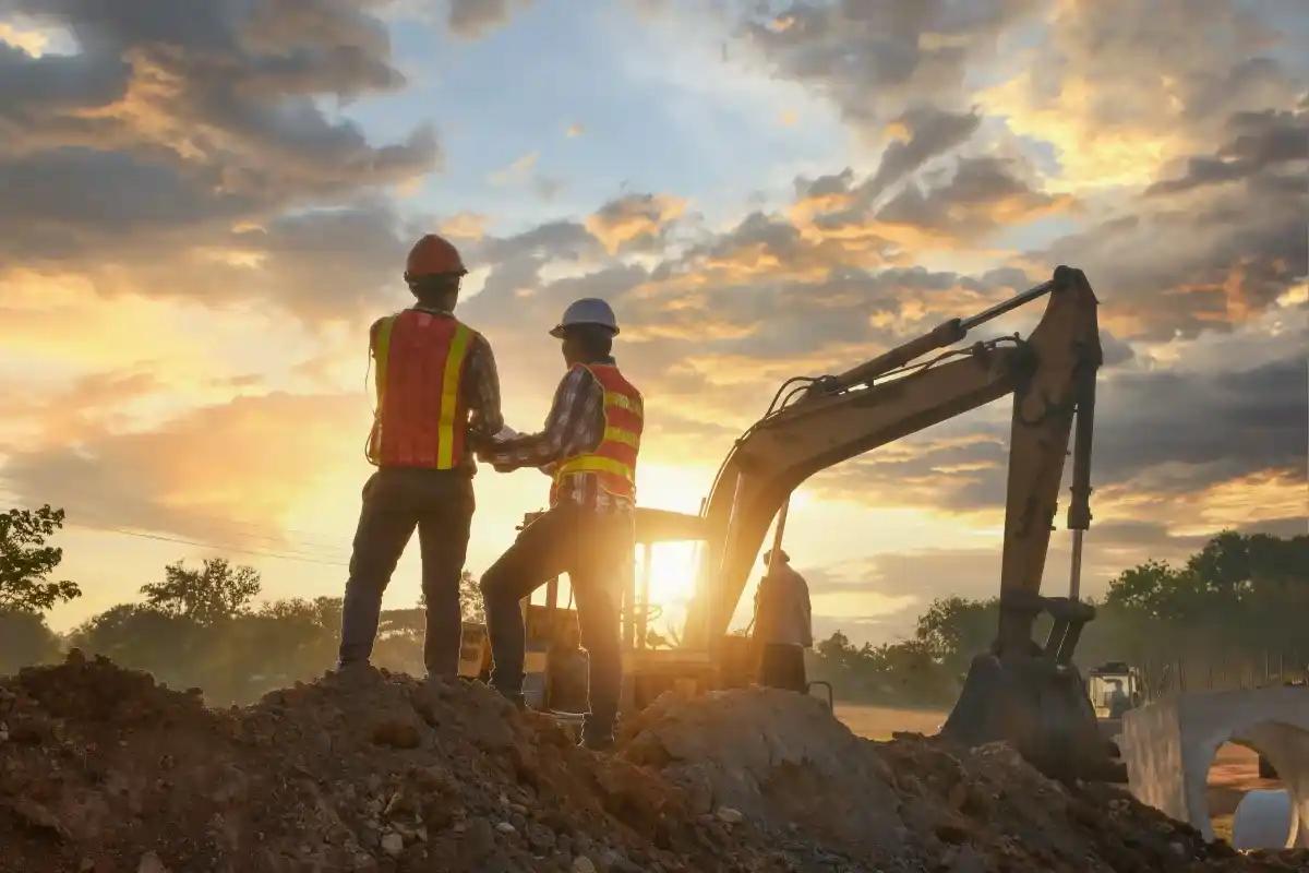 Two construction workers are standing next to an excavator at a construction site at sunset
