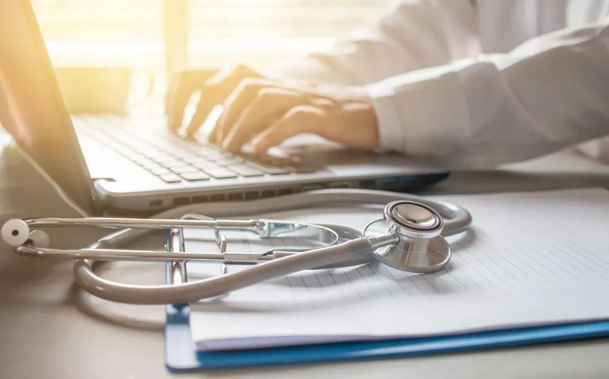 A doctor is typing on a laptop next to a stethoscope and a clipboard