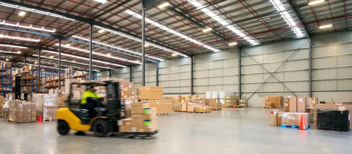 A man is driving a forklift in a large warehouse filled with boxes
