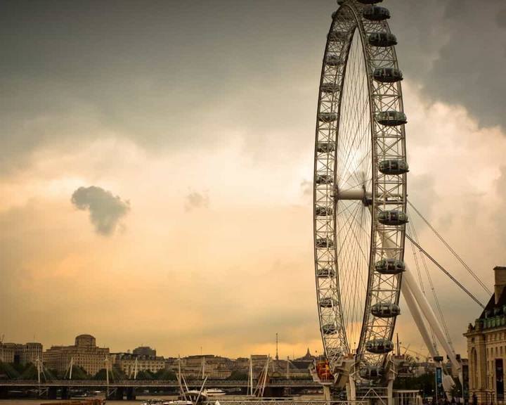 A roda gigante London Eye vista da outra margem do Rio Tâmisa ao anoitecer, com barcos atracados no cais e o céu alaranjado atrás das cabines de vidro.