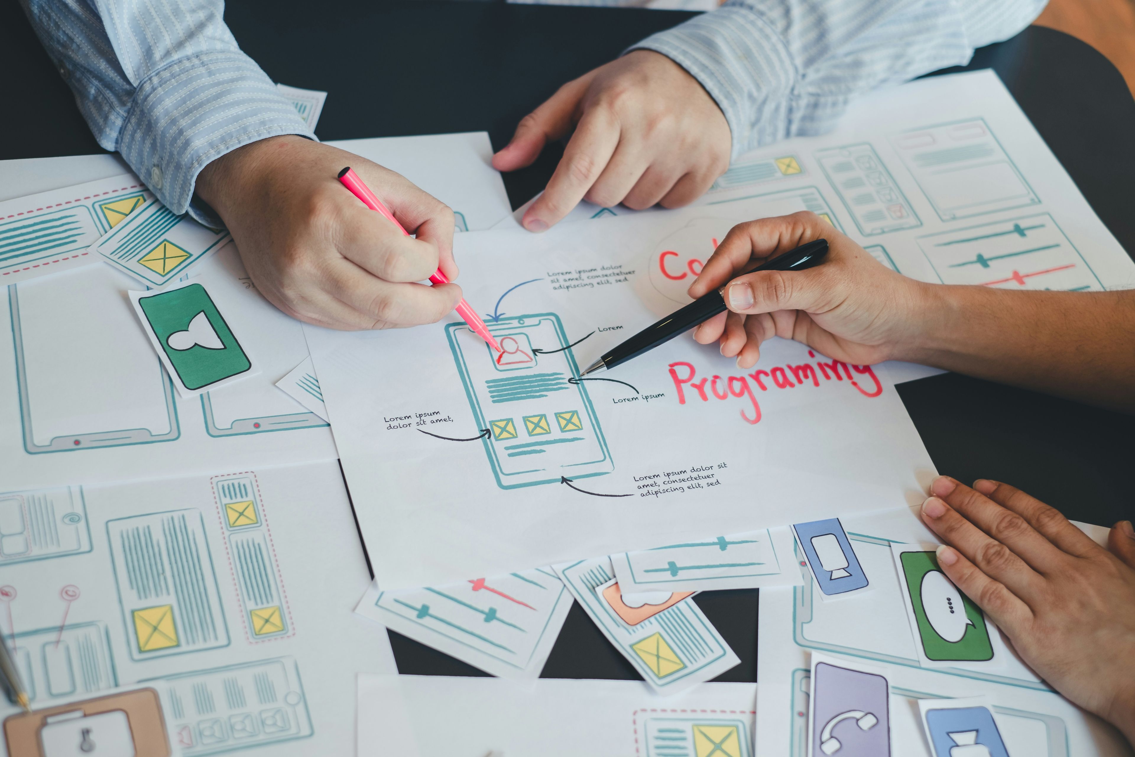 Two designers pointing pens at a paper wireframe laying on a table full of paper wireframes