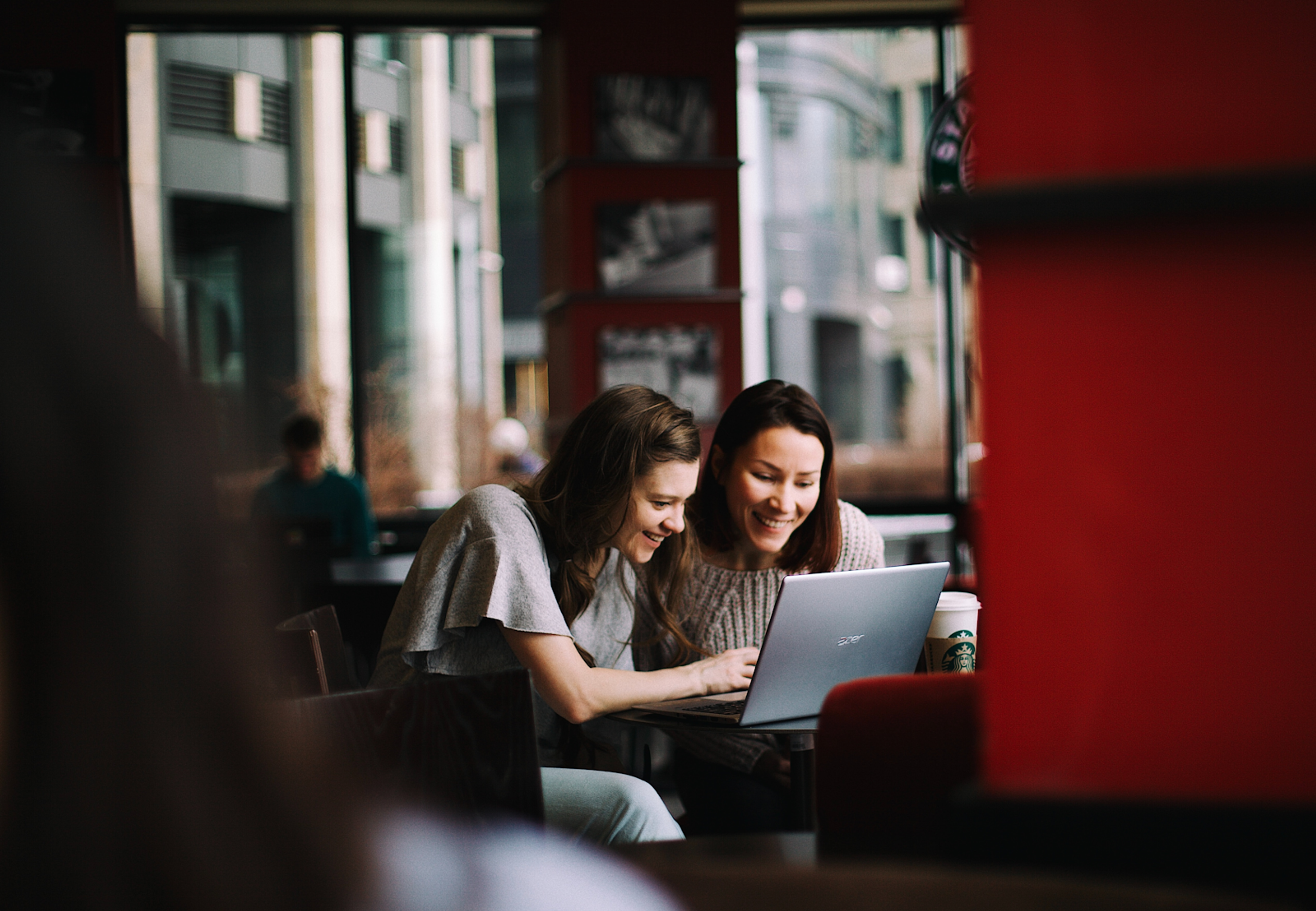 Two women smiling and working together on a laptop in a cozy café setting