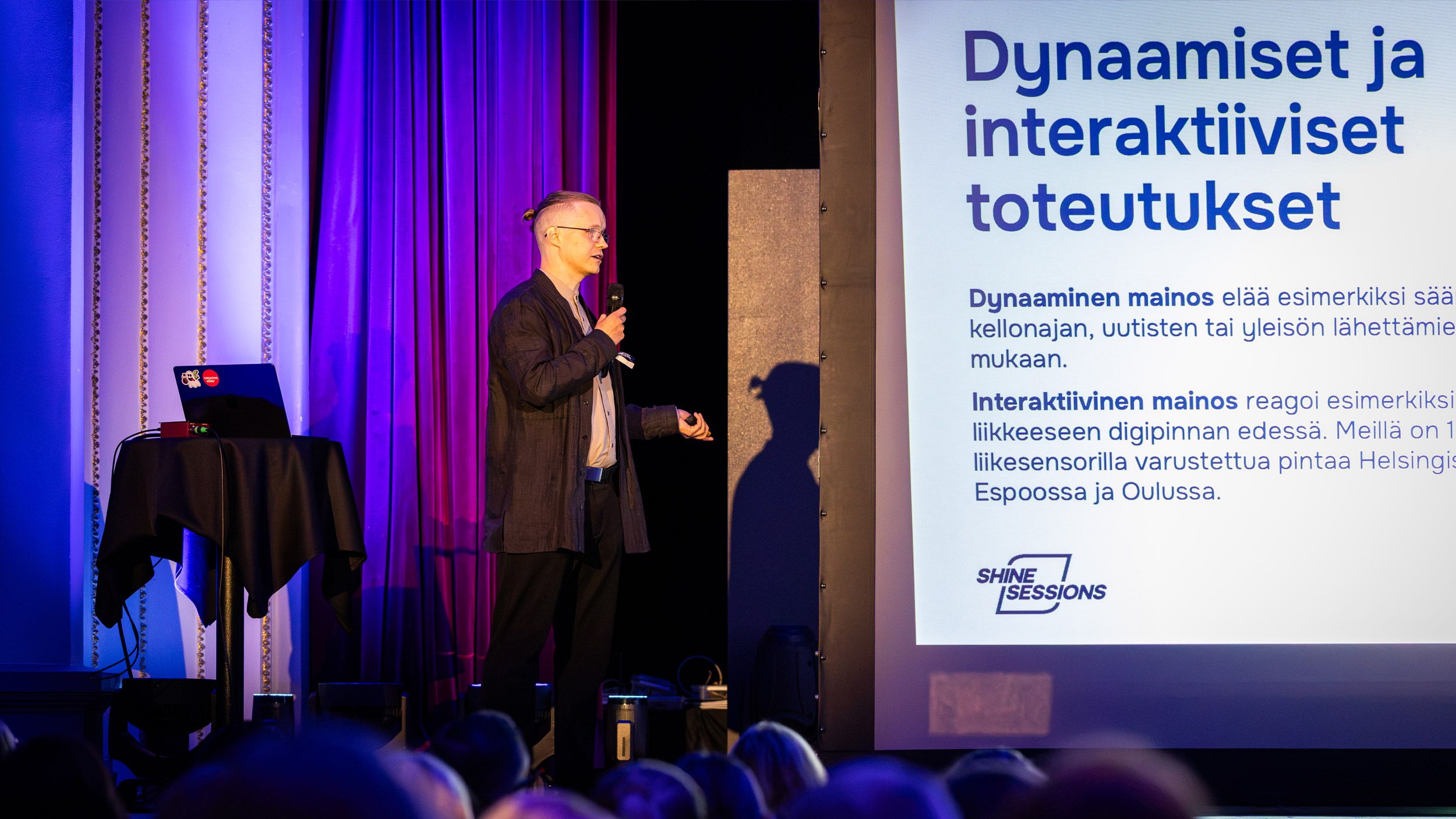 Ilkka Helo kertomassa lavalla mikrofoni kädessä dynaamisista ja interaktiivisista toteutuksista Shine Sessions -tapahtumassa.