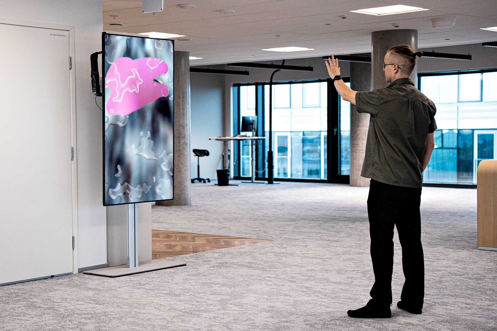 Henkilö ohjaa pystymallista digitaalista näyttöä käden eleillä toimistoympäristössä.