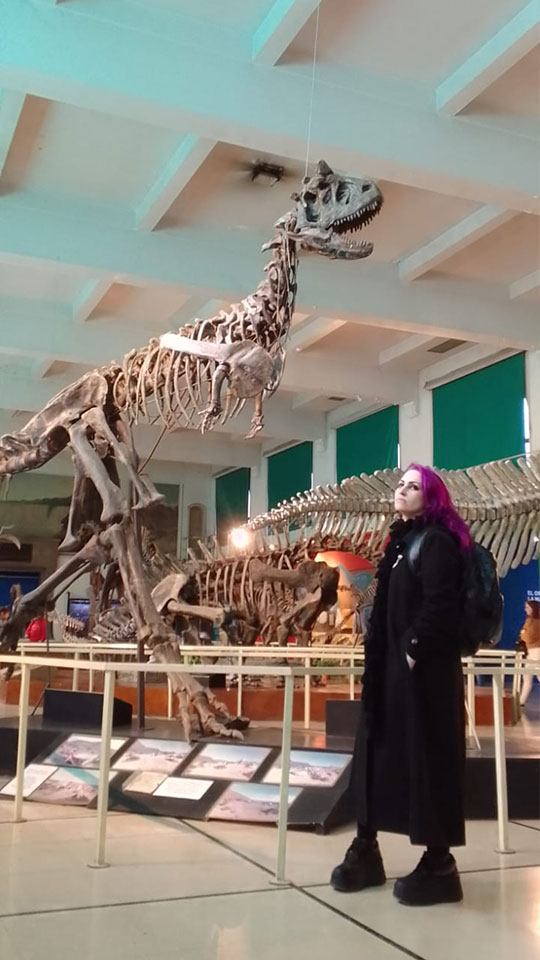 a young woman in a museum posing in front of a Carnotaurus skeleton