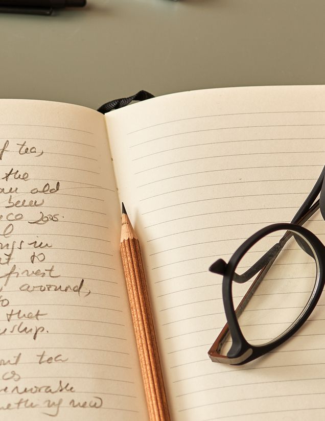 An open notebook with a pencil resting in the fold next to a pair of glasses