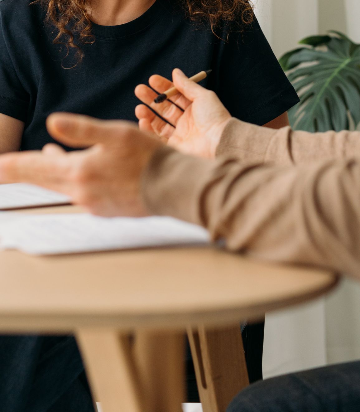 A close up of a pair of hands, open in expression and holding a pen, with a client listening in the background.