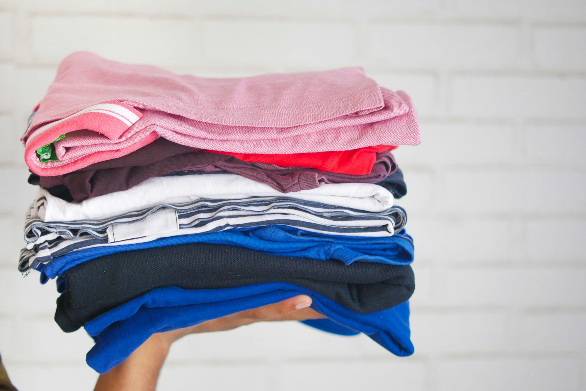 A hand holding a neatly folded stack of pink, blue, white and navy garments against a white wall