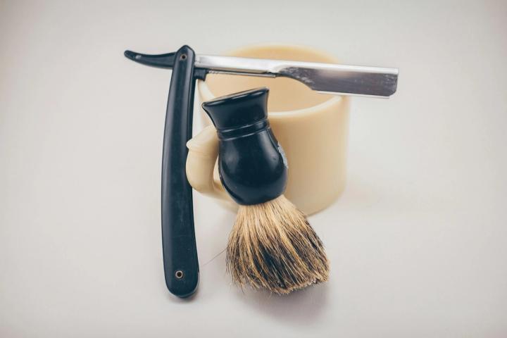 A black straight razor and black-handled shaving brush leaning against a cream ceramic lather mug on a plain light background
