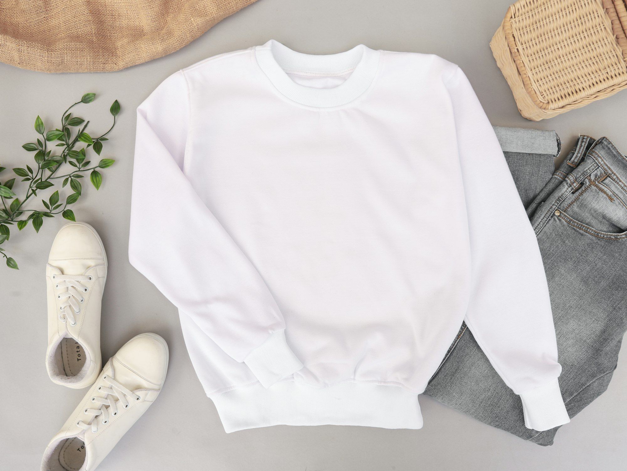 A plain white sweatshirt photographed flat from above alongside jeans, sneakers and a woven basket