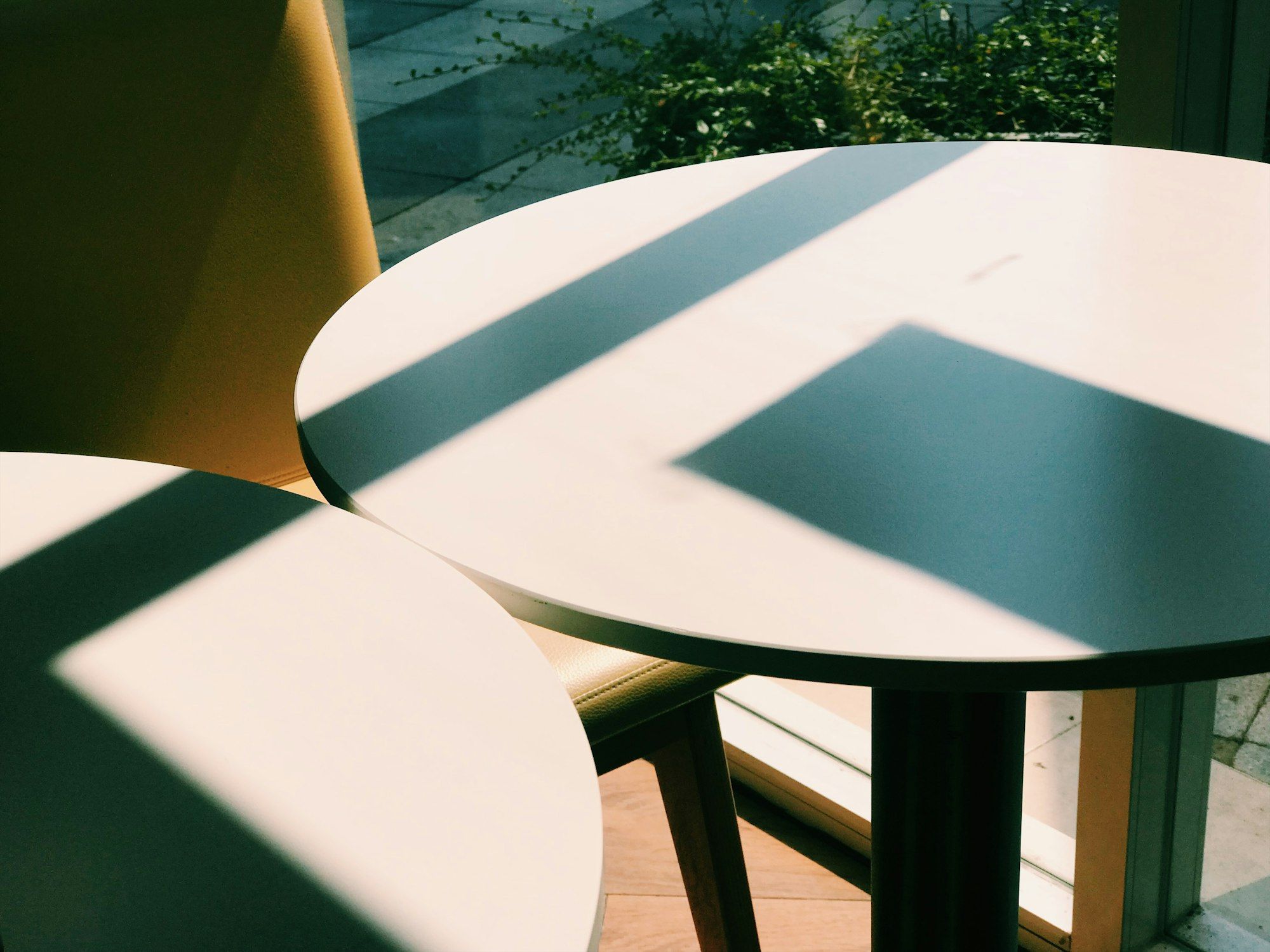 Hard afternoon window light throwing crisp geometric shadow shapes across two round cafe tables.
