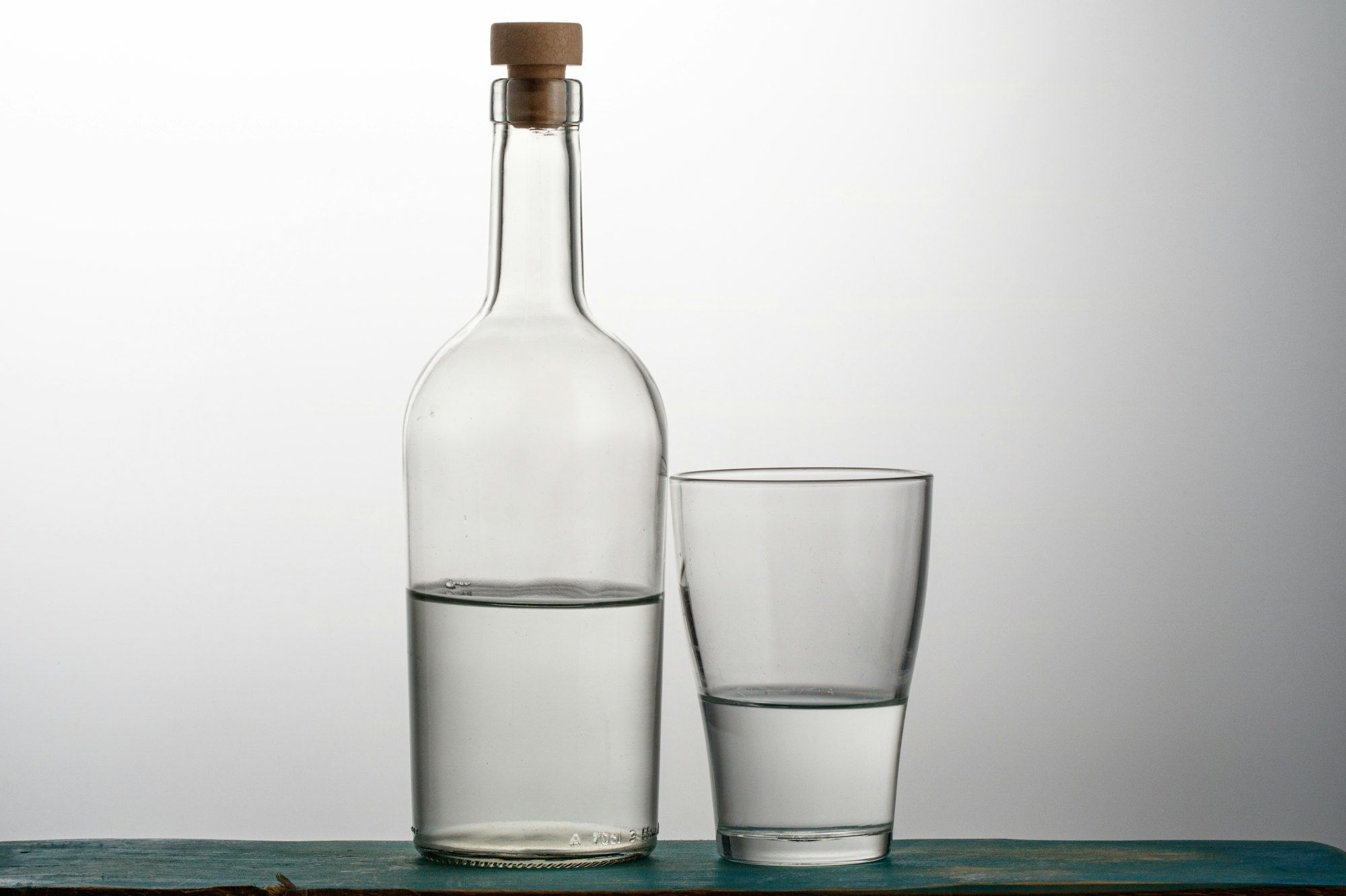 A clear glass bottle with a cork stopper next to a drinking glass, both half full of water, lit against a graduated grey studio background
