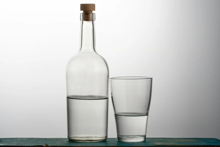 A clear glass bottle with a cork stopper next to a drinking glass, both half full of water, lit against a graduated grey studio background