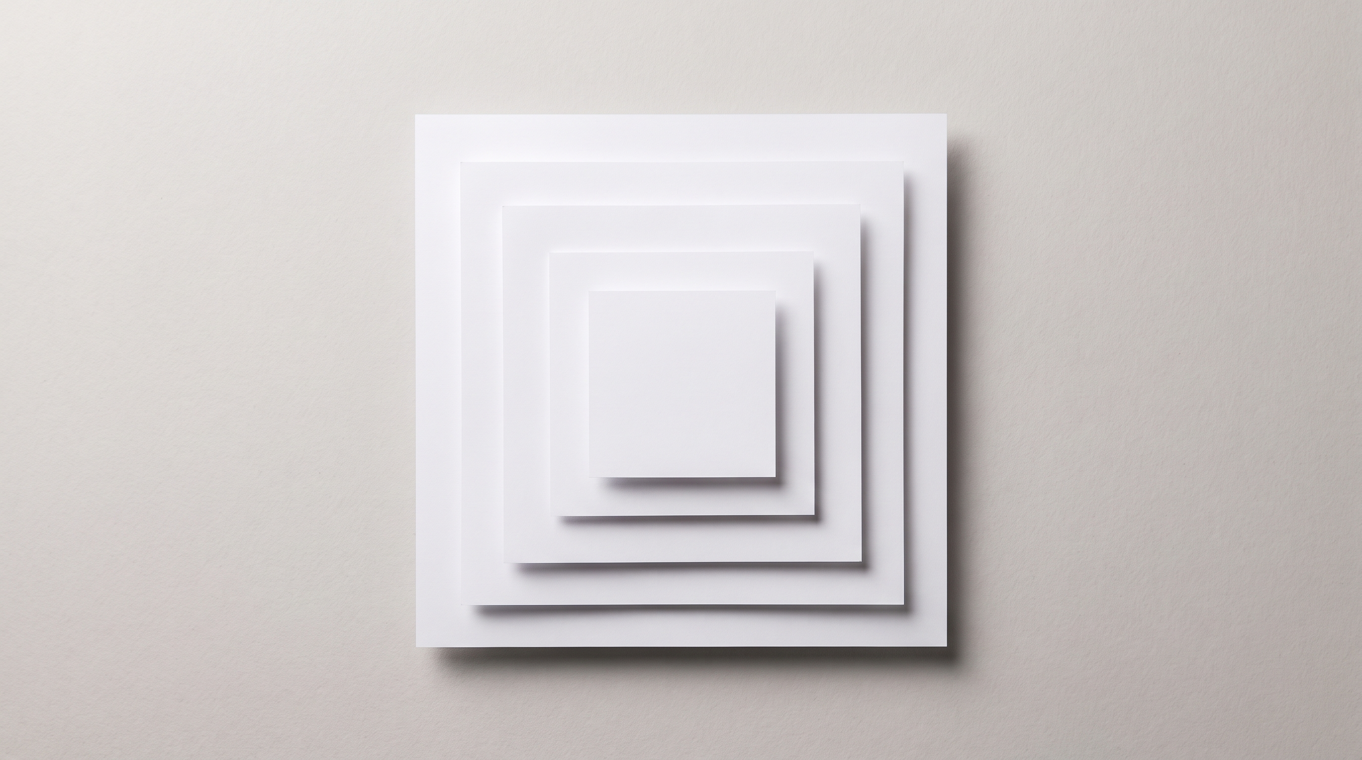 Five plain white paper squares of decreasing size stacked concentrically on a light grey surface