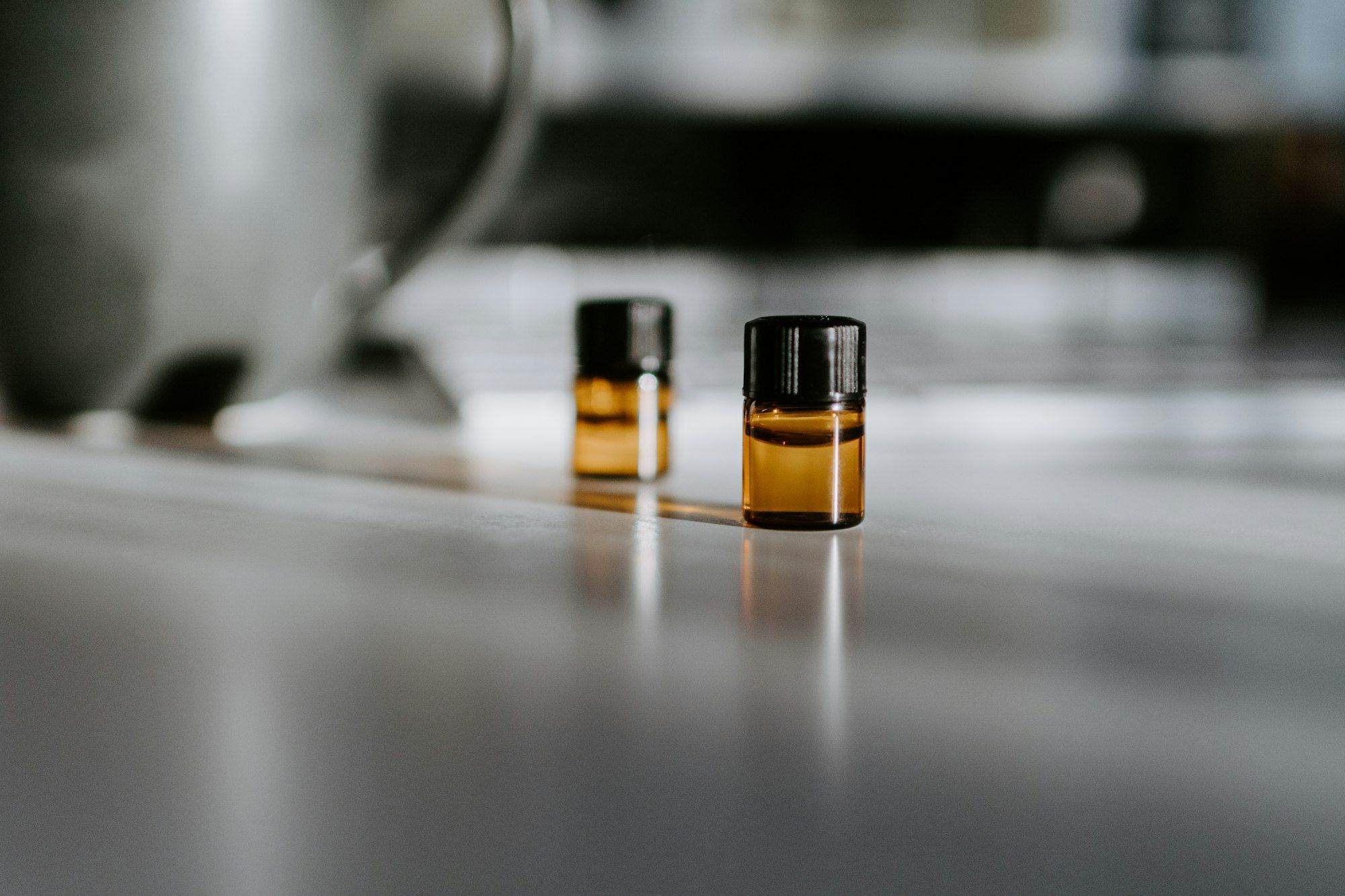 Two small amber glass vials with black caps on a reflective grey surface, each throwing a highlight and a soft reflection