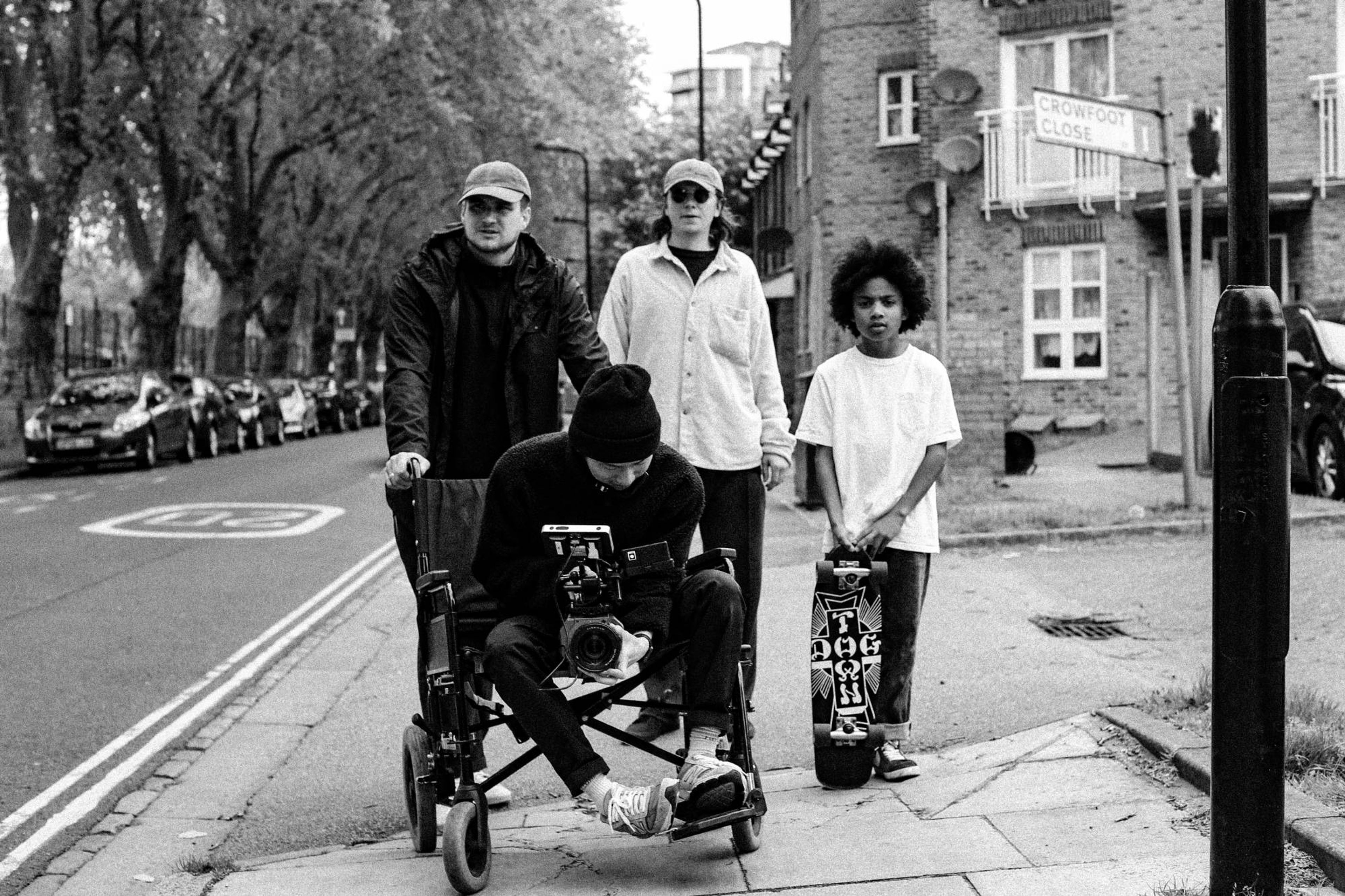 Crew with a camera in a wheelchair on a street corner beside a boy holding a skateboard