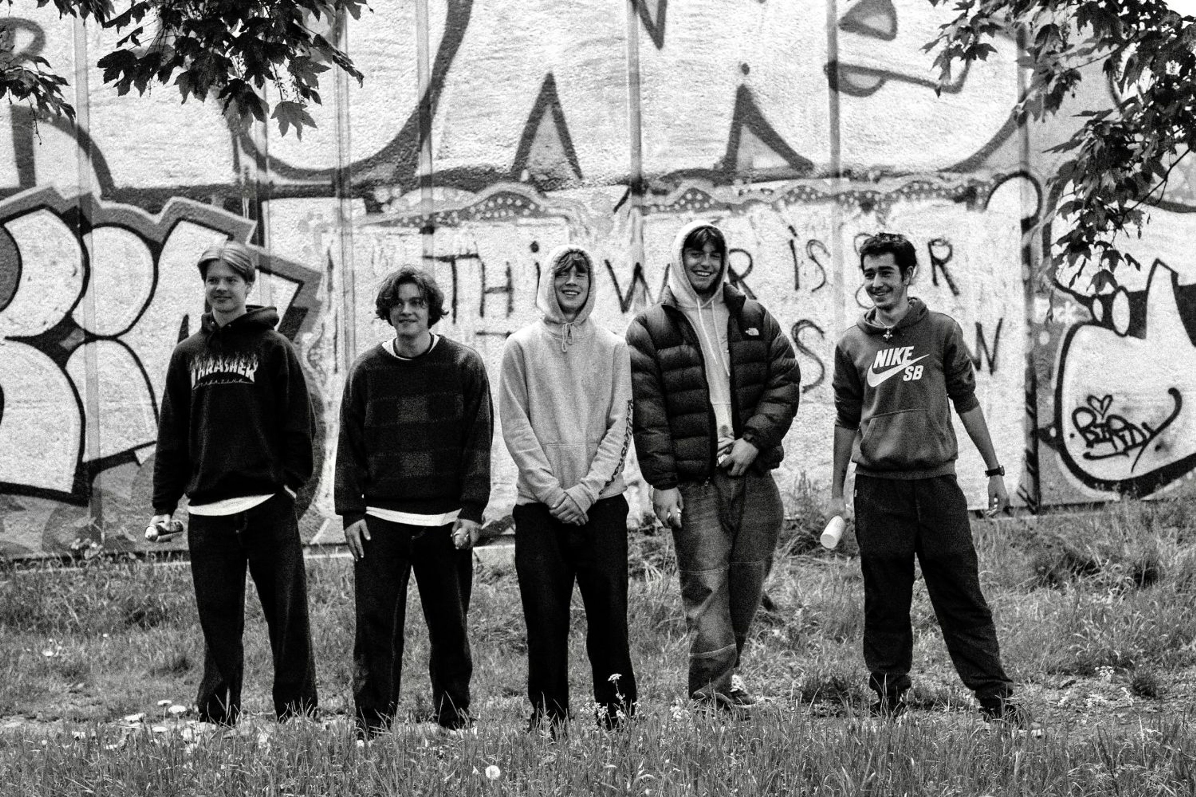 Five skaters smiling in front of a graffitied wall