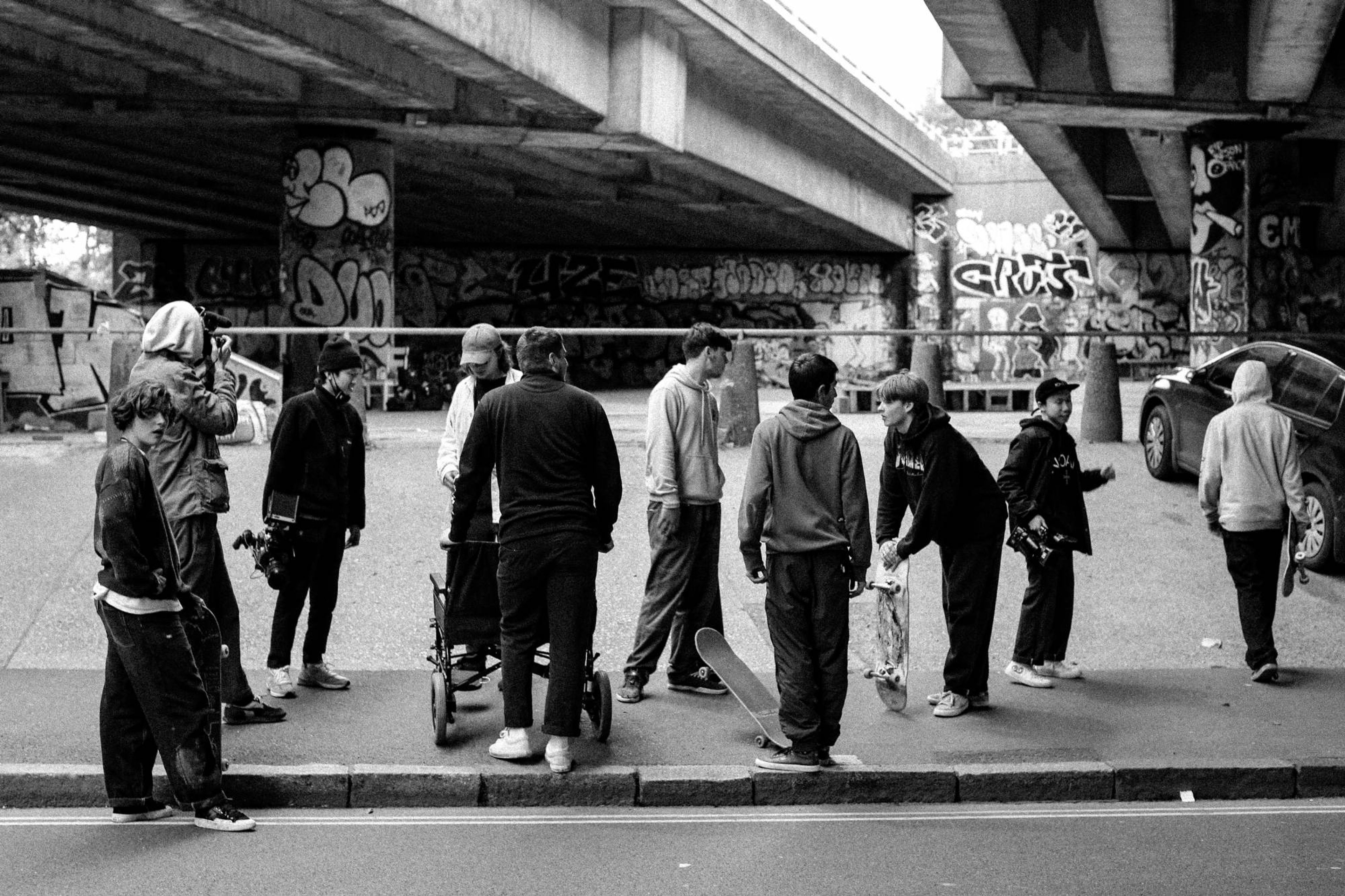 Cast and crew gathered on a pavement under a graffitied flyover