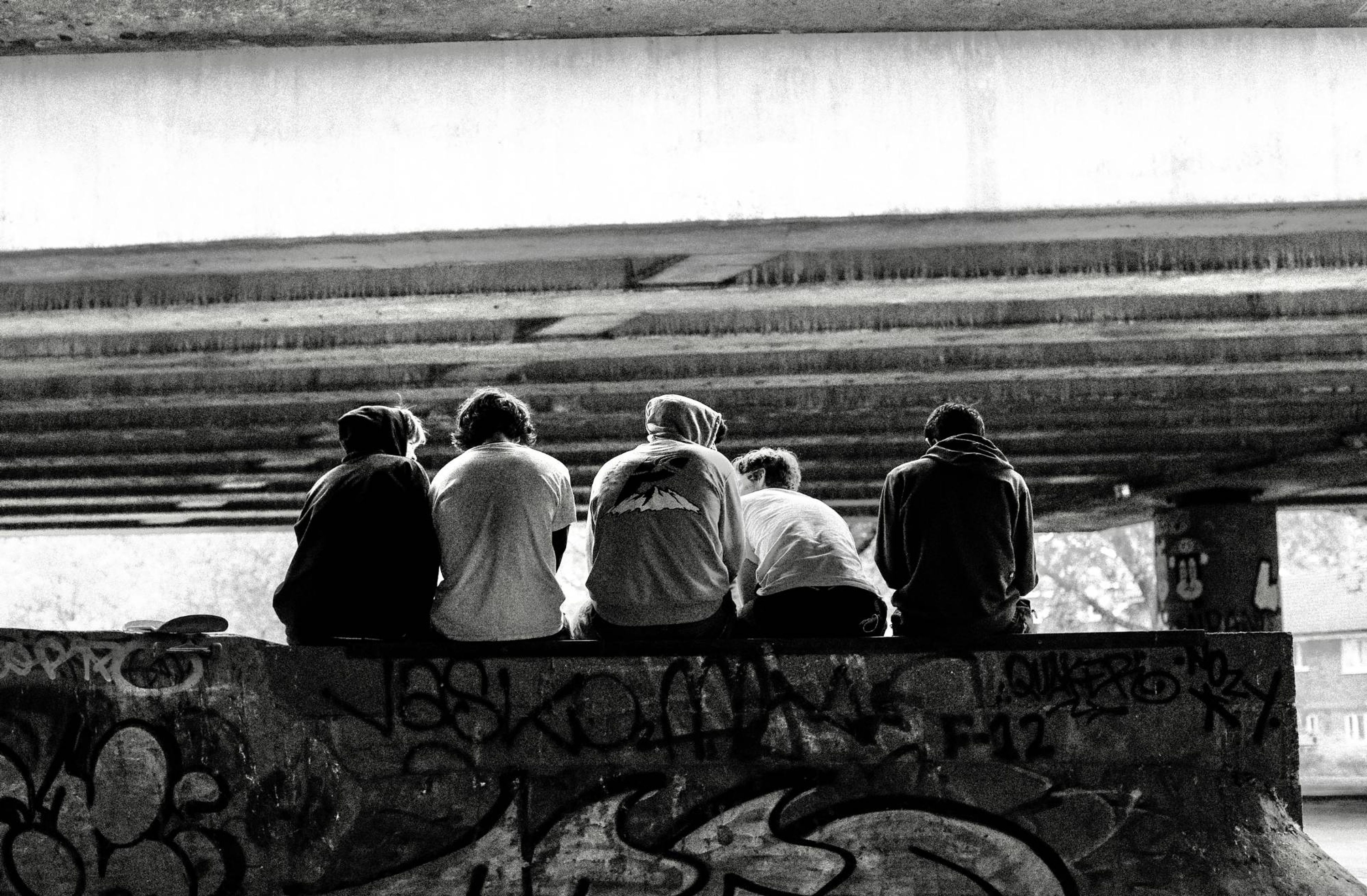 Five skaters sitting on a graffitied wall under a flyover, seen from behind