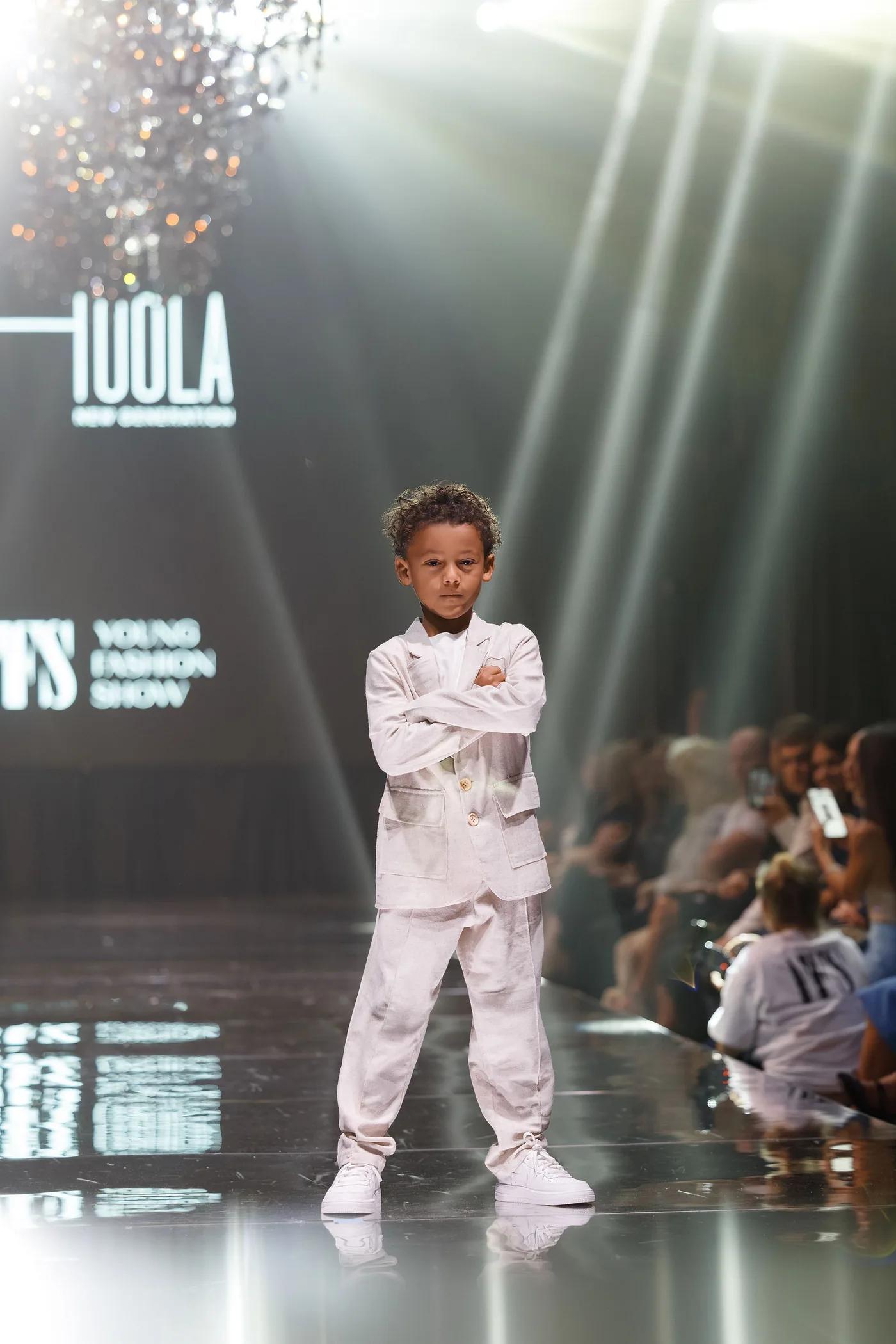 A boy in a white HOOLA suit on the YFS runway, the brand on the screen behind